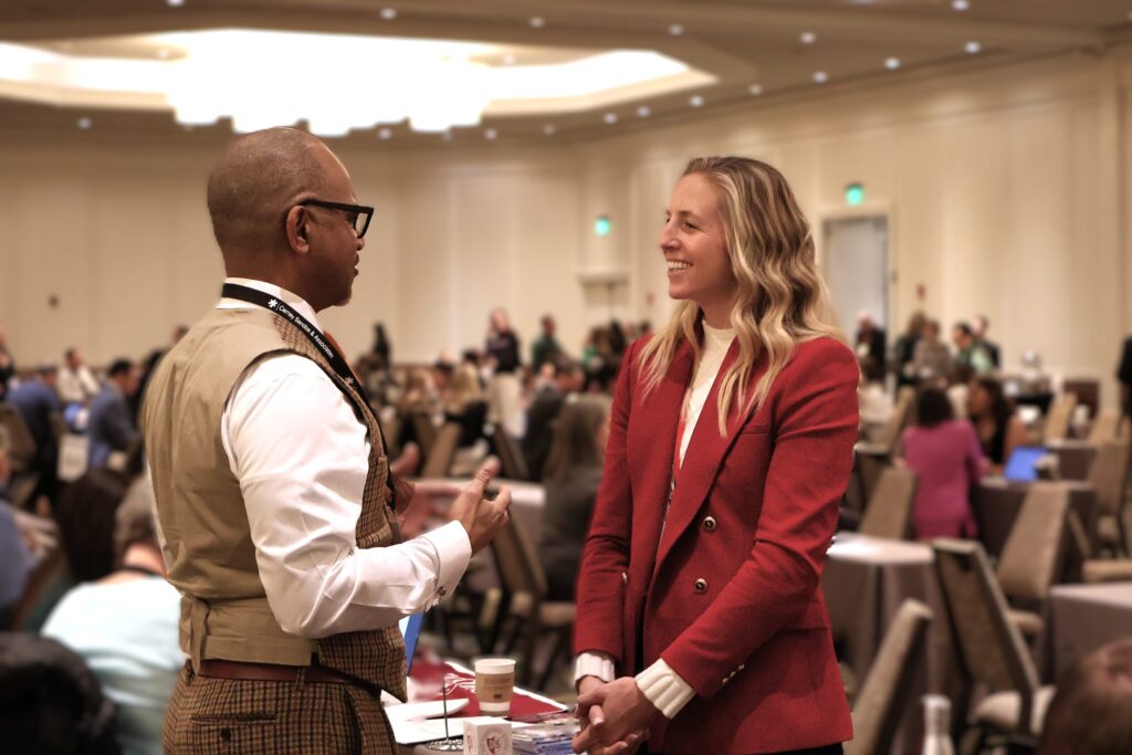 Two people engaged in conversation at a conference with a busy background.